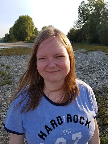 Portrait einer jungen Frau mit langen rötlichen Haaren und einem T-Shirt mit em Aufdruck "Hard Rock". Im Hintegrund ein Kiesstrand. 