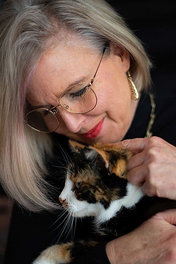 Ein Bild von Petra Pawel, einer Dame in einem schwarzen Rollkragenpullover und goldenen Ohrringen. Sie trägt eine Brille mit goldenem Rand und Maskara und roten Lippenstift. Petra schaut nicht in die Kamera sondern auf die Katze in ihren Armen, eine schöne Katze mit schwarzen, orangenen und weißen Stellen. Die Katze sieht glücklich darüber aus gestreichelt zu werden und Petra sieht glücklich darüber aus sie zu streicheln.