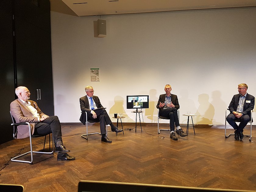 Diskussionsrunde, vier Männer im Anzug auf Stühlen sitzend, in der Mitte steht auf einem Tisch ein Bildschirm, der die zugeschalteten Teilnehmer zeigt.