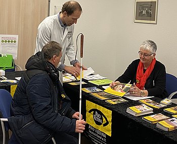 Priv.-Doz. Dr. Boris Stanzel, Leiter des Makulazentrums an der Augenklinik Sulzbach, stehend in der Mitte des Bildes, und Roswitha Karst, PRO RETINA, beraten einen Besucher des AMD-Thementags am Infostand von PRO RETINA.