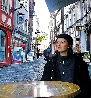 Eine junge Frau sitzt an einem Tisch in der Fußgängezone. Sie trägt einen schwarzen Mantel und eine schwarze Mütze.