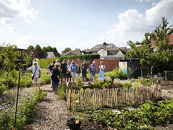 Etwa 12 Teilnehmer stehen im kulturellen Garten. Sie unterhalten sich und schauen sich die Pflanzenbeete an. Im Vordergrund sieht man verschiedene Beete mit Blumen und Gemüse, die von kleinen Holzzäunen umgeben sind. Die Wege sind mit Holzschnitzeln bedeckt. Im Hintergrund stehen einige Wohnhäuser mit Gärten, Hecken und Bäumen. Das Wetter ist sonnig mit ein paar Wolken am Himmel. Die Stimmung wirkt entspannt und freundlich.
