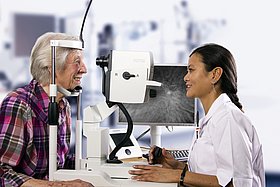 Eine Patientin hat ihr Kinn auf eine OCT Untersuchungsgerät gelegt. Eine Ärtzin fotografiert die Netzhaut.