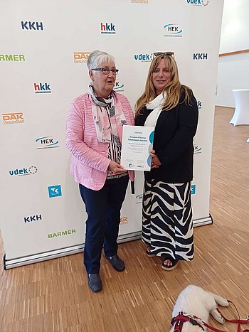 Bild von Roswitha Kaarst (links) und Marion Palm-Stalp (rechts). Sie halten gemeinsam eine Urkunde über den erhaltenen Preis in die Kamera. Zu sehen ist auch rechts in der Ecke ein Hund mit weißen Fell. Hinter den beiden Frauen ist ein RollUp mit den Logos der Ersatzkassen aufgestellt.