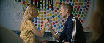 Auf dem Bild zu sehen sind eine Frau mit langen, blonden Haaren in einem gelb gestreiften Shirt und ein Mann mit kurz rasierten Haaren in einer blauen Trainingsjacke. Der Mann sitzt der Frau mit geschlossenen Augen sehr nah gegenüber und hat seine Hand gehoben, die Frau berührt mit ihrem Zeigefinger seine Finger als würde sie etwas zählen