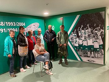 Die Gruppe sitzt vor einer grünen Wand. Links von ihnen ist ein Foto von Fußballspielern zu sehen. Die Fußballspieler jubeln. Einer streckt einen Pokal in die Luft
