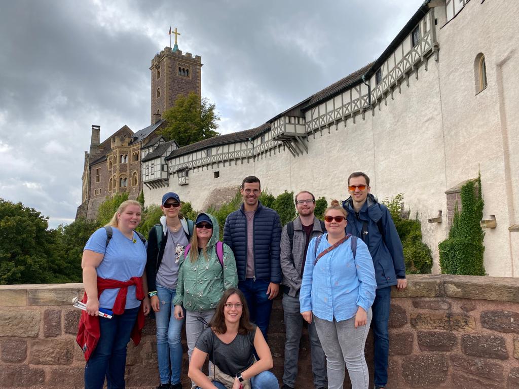 Auf dem Foto sind die acht Teilnehmenden des Summer Mettups zu sehen. Sie stehen vor einer Mauer und im Hintergrund ist die Wartburg zu sehen. Eine Frau kniet im Vordergrund und die anderen stehen hinter ihr. Manche tragen Sonnenbrillen und haben ihren zusammengeklappten Langstock in der Hand.